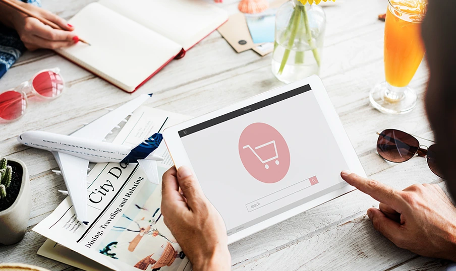 Person uses tablet with shopping cart icon, surrounded by travel magazines, airplane, and drinks.